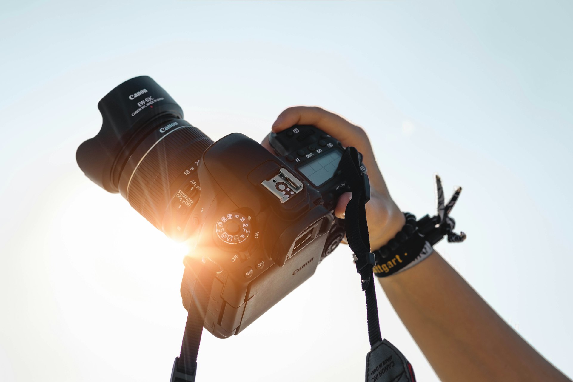 A hand raising a camera toward a bright sky with a warm sun flare